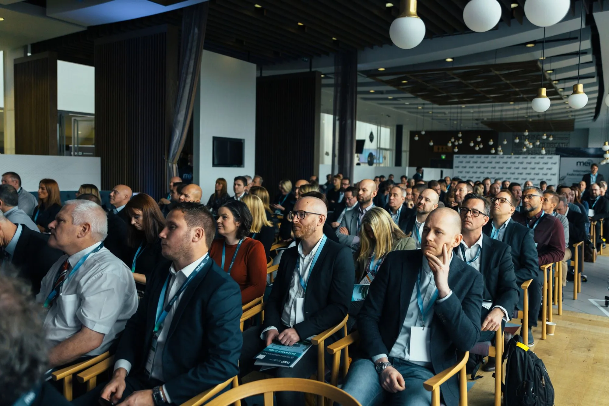 A packed conference audience wearing lanyards, attentively listening in a bright modern venue
